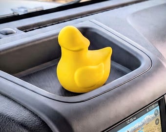May include: A bright yellow rubber duck figurine with a smooth, rounded body and a distinct beak, positioned inside the recessed storage tray of a vehicle dashboard. The duck is solid in color and features a simple, stylized avian silhouette.