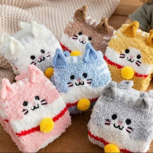 May include: Six pairs of fluffy cat-themed socks in various colours, including pink, blue, white, brown, and yellow. Each sock features a cat face with embroidered details and a fluffy pom-pom.