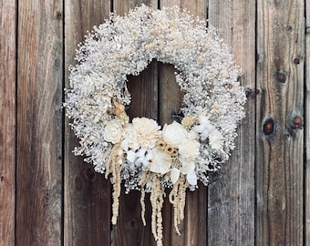 May include: A white dried flower wreath with white and cream colored flowers and greenery. The wreath is hanging on a wooden wall.
