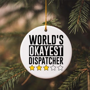 Könnte beinhalten: Weißer Keramikschmuck mit dem schwarzen Text "WORLD'S OKAYEST DISPATCHER". Der Schmuck hat drei gelbe Sterne und zwei weiße Sterne. Er hängt an einer goldenen Schnur vor einem Hintergrund aus grünen Tannennadeln.