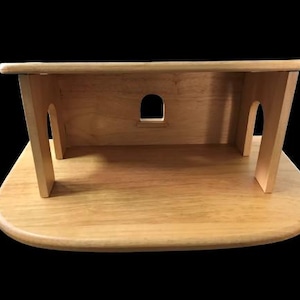 Può includere: Una casetta di legno con base arrotondata e tetto piatto. La casetta ha due aperture ad arco e una finestra rettangolare sul retro.