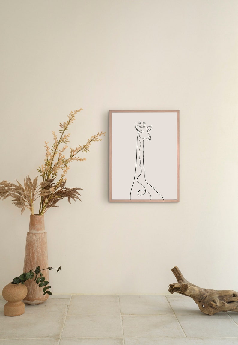 May include: A black and white line drawing of a giraffe in a simple, minimalist style. The drawing is framed in a light brown frame and is hanging on a white wall. A vase with dried flowers and a piece of driftwood are in the foreground.