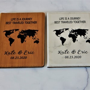 May include: Two passport covers with a world map design. One cover is brown with a wood grain texture and the other is white with a marble texture. Both covers have the text "Life is a journey best traveled together" and "Kate & Eric 08.23.2020" engraved on them.