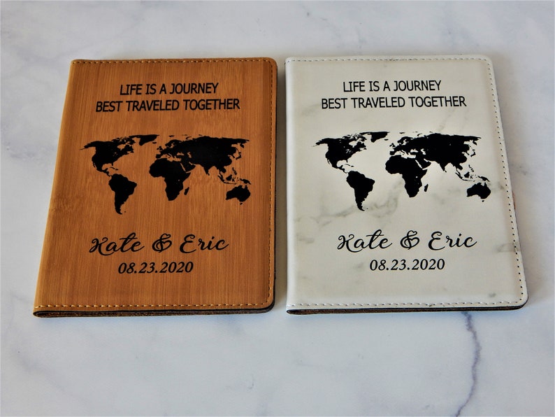 May include: Two passport covers with a world map design. One cover is brown with a wood grain texture and the other is white with a marble texture. Both covers have the text "Life is a journey best traveled together" and "Kate & Eric 08.23.2020" engraved on them.