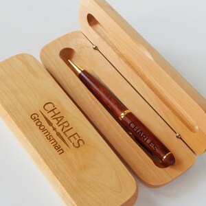 May include: A wooden pen and case set. The pen is dark brown with gold accents and the name "CHARLES" engraved on it. The light-colored wooden case is open, revealing the pen, and also has the name "CHARLES" and the word "Groomsman" engraved on it.