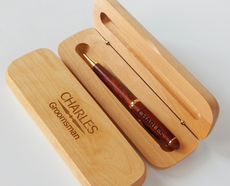 May include: A wooden pen and case set. The pen is dark brown with gold accents and the name "CHARLES" engraved on it. The light-colored wooden case is open, revealing the pen, and also has the name "CHARLES" and the word "Groomsman" engraved on it.