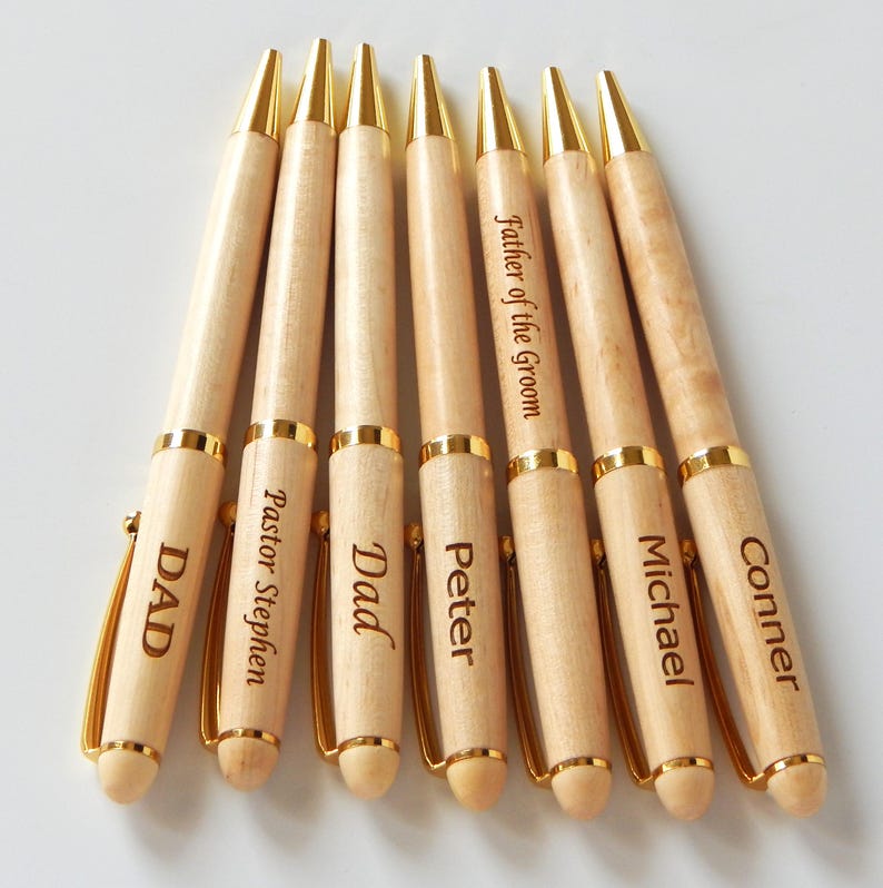 May include: Seven wooden pens with gold accents and personalized inscriptions. The pens are light brown with gold tips, clips, and bands. Each pen has a different name or title engraved on it, such as "DAD" and "Father of the Groom".