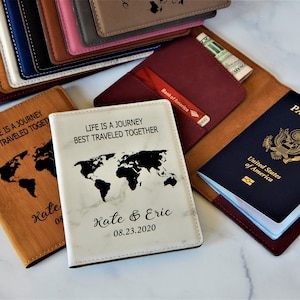 May include: Two passport covers with a world map design and the text "Life is a journey best traveled together". One cover is brown with the text "Kate 08" and the other is white with the text "Kate & Eric 08.23.2020".
