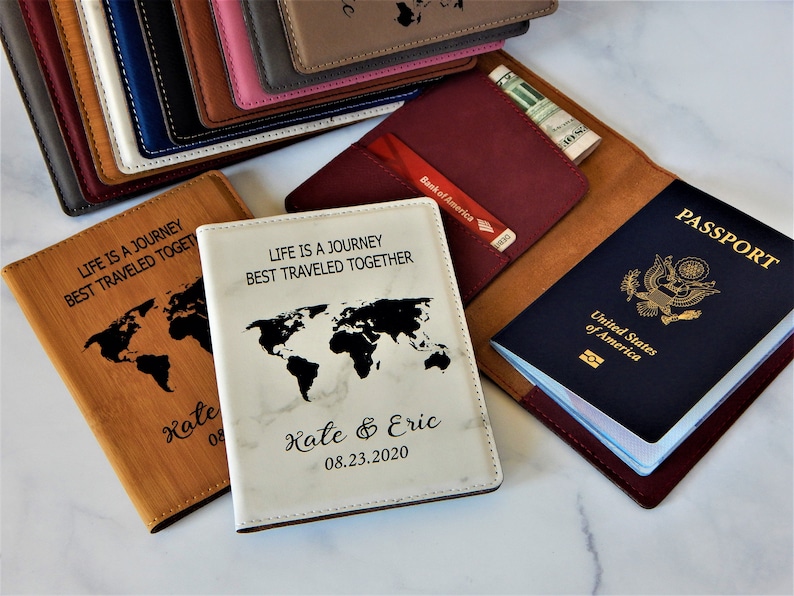 May include: Two passport covers with a world map design and the text "Life is a journey best traveled together". One cover is brown with the text "Kate 08" and the other is white with the text "Kate & Eric 08.23.2020".