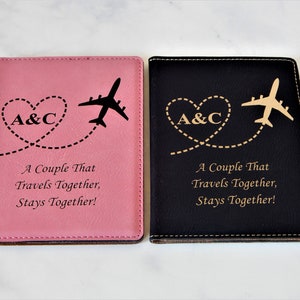May include: Two passport holders, one pink and one black, with a gold airplane and heart design. The text "A Couple That Travels Together, Stays Together!" is engraved on both holders.