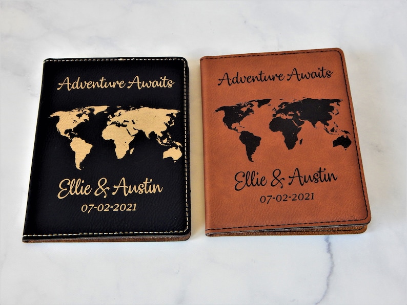 May include: Two passport covers with a world map design. One cover is black with a gold world map and the other is brown with a black world map. Both covers have the text "Adventure Awaits" and "Ellie & Austin 07-02-2021" engraved on them.