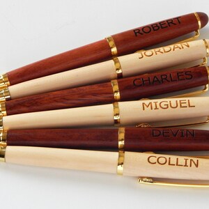 May include: Six personalized wooden pens with gold-colored accents. The pens are engraved with names: ROBERT, JORDAN, CHARLES, MIGUEL, DEVIN, and COLLIN. The pens alternate between a dark brown and a light wood color.