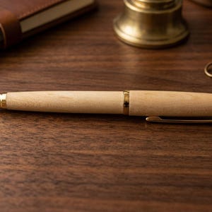 May include: A ballpoint pen featuring a light-colored wooden barrel with a smooth finish, accented by polished gold-tone metal components including the tip, center band, pocket clip, and end cap, presented in a sleek, cylindrical design.
