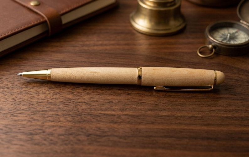 May include: A ballpoint pen featuring a light-colored wooden barrel with a smooth finish, accented by polished gold-tone metal components including the tip, center band, pocket clip, and end cap, presented in a sleek, cylindrical design.