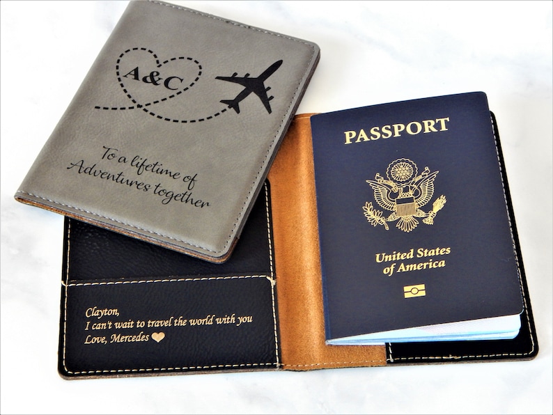 May include: A gray and black passport holder with a brown leather trim. The front cover has a heart with the initials "A&C" and a plane with a dotted line trail. The text "To a lifetime of Adventures together" is engraved on the front cover. The back cover has the text "Clayton, I can't wait to travel the world with you Love, Mercedes".