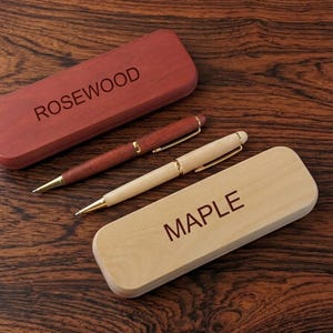 May include: A dark reddish-brown rosewood pen and matching case with the word ROSEWOOD engraved on the lid, alongside a light tan maple pen and matching case with the word MAPLE engraved on the lid, both featuring gold-toned metal accents.