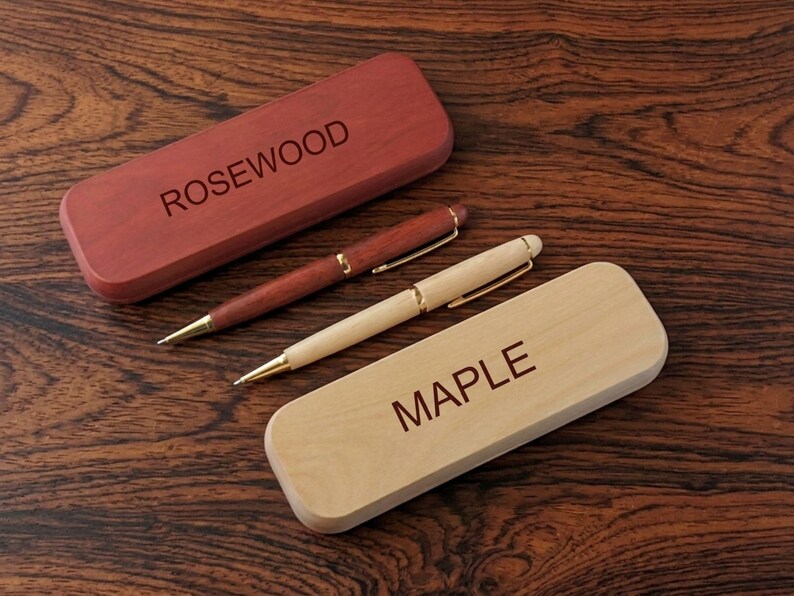 May include: A dark reddish-brown rosewood pen and matching case with the word ROSEWOOD engraved on the lid, alongside a light tan maple pen and matching case with the word MAPLE engraved on the lid, both featuring gold-toned metal accents.