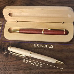 May include: A dark reddish-brown wooden pen with gold-tone accents rests inside a light-colored wooden case measuring 6.5 inches by 2.0 inches. A second light-colored wooden pen with gold-tone accents measures 5.5 inches in length.