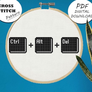 May include: A white cross stitch pattern in a wooden embroidery hoop with black text that reads "Ctrl + Alt + Del". The hoop is on a blue background with a potted plant to the right.