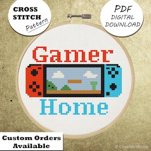 May include: A cross-stitch pattern in a wooden hoop. The design features the word "Gamer" in red, a pixelated video game controller, and the word "Home" in blue. The text "Cross Stitch Pattern", "PDF Digital Download", and "Custom Orders Available" are also present.