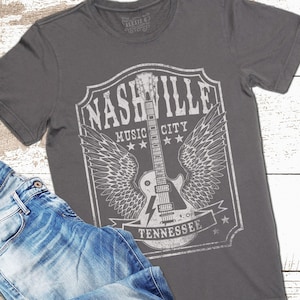 May include: A charcoal grey t-shirt featuring a vintage-style graphic of a guitar with wings. The graphic reads "Nashville Music City Tennessee" and has a distressed look.