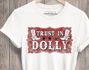 May include: White t-shirt with a red and white graphic that reads "TRUST IN DOLLY". The design includes two cowboy boots and the text "DOLLY" in large, bold letters. The shirt is hanging on a wooden hanger.