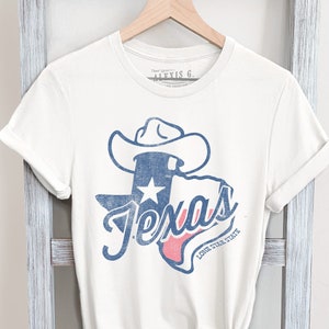 May include: A white t-shirt with a blue and red graphic of the state of Texas with a cowboy hat on top. The graphic says "Texas" and "Lone Star State".