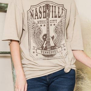May include: A beige t-shirt featuring a graphic print of a guitar with wings and the text "Nashville Music City Tennessee".