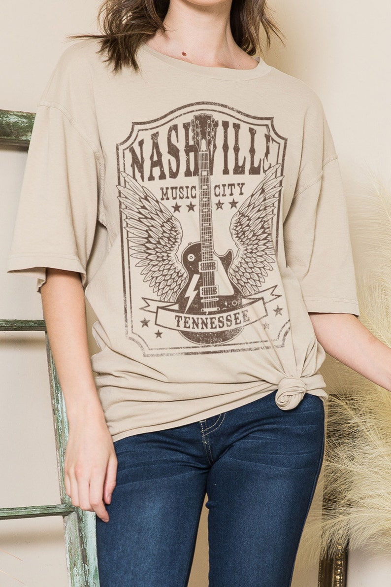 May include: A beige t-shirt featuring a graphic print of a guitar with wings and the text "Nashville Music City Tennessee".