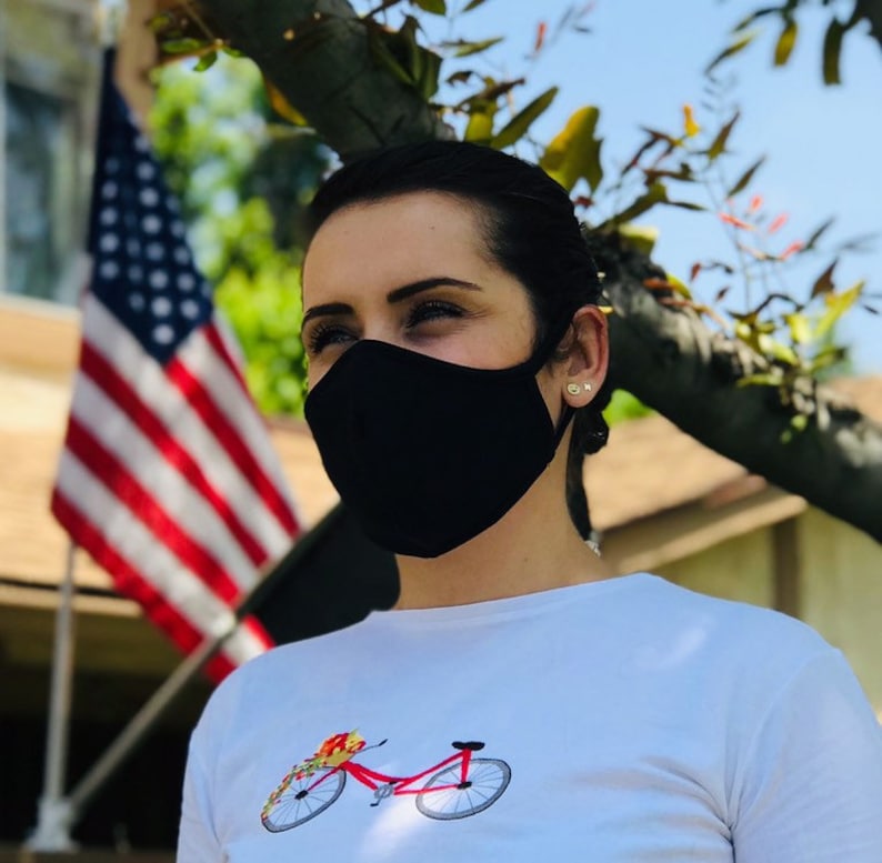 Op de afbeelding: Een vrouw draagt ​​een zwart mondkapje en een wit T-shirt met een rode fietsafbeelding. De fiets heeft een mand vol bloemen.