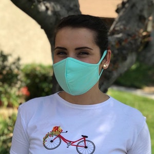 Op de afbeelding: Een persoon draagt ​​een lichtblauw mondkapje met een wit T-shirt met daarop een rode fiets met een mand vol bloemen.