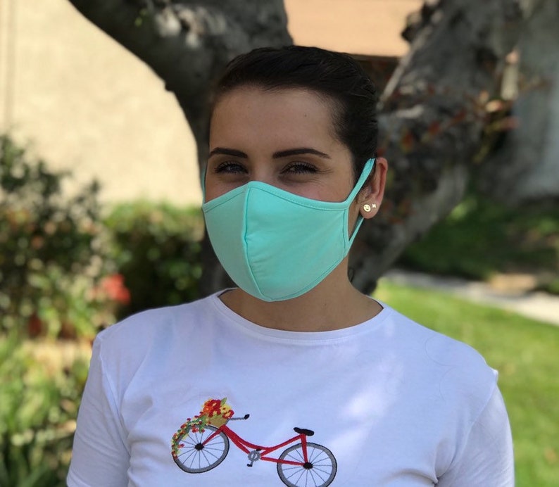 Op de afbeelding: Een persoon draagt ​​een lichtblauw mondkapje met een wit T-shirt met daarop een rode fiets met een mand vol bloemen.