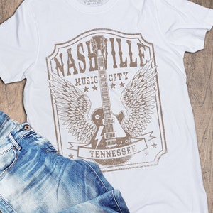 May include: White t-shirt featuring a brown graphic print of a guitar with wings and the text "Nashville Music City Tennessee".