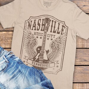 May include: A beige t-shirt featuring a vintage-style graphic of a guitar with wings and the text "Nashville Music City Tennessee".