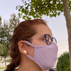 Op de afbeelding: Een persoon draagt ​​een lichtpaarse gezichtsmasker met een bril met blauwe monturen. De persoon draagt ​​een zwart shirt en heeft bruin haar.