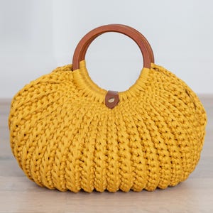 May include: A mustard yellow crocheted handbag featuring a rounded, ribbed texture and a circular brown wooden handle. The bag includes a small brown leather-like tab closure at the top center, securing the opening between the two wooden handles.