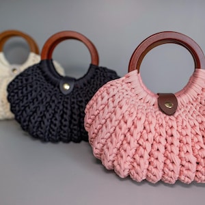 May include: Three crescent-shaped handbags made of thick, textured knit fabric in cream, black, and dusty pink. Each bag features a round, dark brown wooden handle and a small, centered tab closure with a metallic snap button at the top opening.