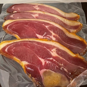 May include: Four slices of cured meat, likely ham or prosciutto, are vacuum-sealed in clear plastic. The meat is a deep red color with a lighter, yellowish-brown fat cap around the edges. The packaging is slightly reflective, and the meat appears fresh.