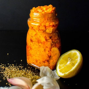 May include: A glass jar filled with a bright orange, chunky condiment. The jar is sitting on a black surface with a lemon wedge, garlic cloves, and dried herbs.