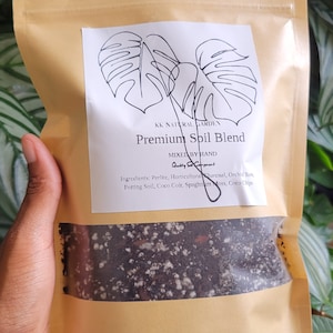 May include: A brown paper bag with a white label that says "KK Natural Garden Premium Soil Blend Mixed by Hand Quality Soil Components" and lists the ingredients: Perlite, Horticultural Charcoal, Orchid Bark, Potting Soil, Coco Coir, Spaghnum Moss, Coco Chips. The bag is filled with a dark brown soil mix.