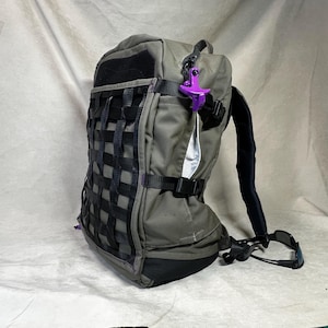 Peut inclure: Un sac à dos tactique vert olive doté d'une grille noire de sangles sur le devant, d'une base noire et d'un clip en plastique violet attaché à la fermeture éclair latérale. Le sac comprend des bretelles noires et une boucle de compression.