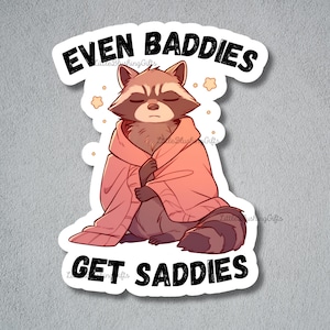 K&ouml;nnte beinhalten: Ein Aufkleber mit einem Cartoon-Waschb&auml;ren, der einen rosa Pullover tr&auml;gt, mit dem Text "Even Baddies Get Saddies".