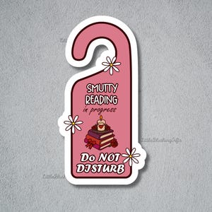 Puede incluir: Una pegatina rosa con forma de colgador de puerta con el texto "Smutty Reading in Progress" y "Do Not Disturb". La pegatina presenta una pila de libros, una vela encendida y detalles de margaritas.