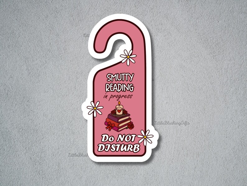 Puede incluir: Una pegatina rosa con forma de colgador de puerta con el texto "Smutty Reading in Progress" y "Do Not Disturb". La pegatina presenta una pila de libros, una vela encendida y detalles de margaritas.