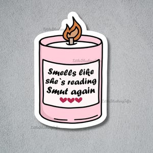 Puede incluir: Una pegatina de vela rosa con una llama encendida. La pegatina tiene el texto "Smells like she's reading Smut again" con tres corazones debajo. Fondo gris.