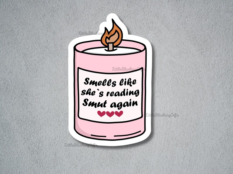 Puede incluir: Una pegatina de vela rosa con una llama encendida. La pegatina tiene el texto "Smells like she's reading Smut again" con tres corazones debajo. Fondo gris.
