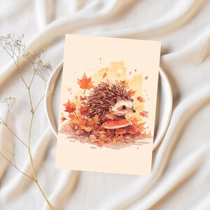 K&ouml;nnte beinhalten: Eine Aquarell-Illustration eines Igels mit braunem und wei&szlig;em Fell, der auf einem Pilz in einer Waldlandschaft sitzt. Der Igel ist von Herbstbl&auml;ttern in Orange-, Rot- und Gelbt&ouml;nen umgeben.