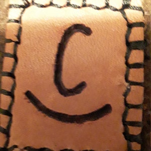 May include: A brown leather patch with black stitching around the edges. The patch has a black letter "C" and a black smiling mouth.