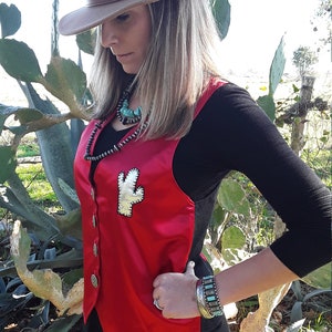 May include: A tan fedora hat with a brown leather band and buckle. A red vest with a cactus patch and silver buttons is worn over a black long-sleeve shirt. The person is wearing blue jeans and a turquoise bracelet.