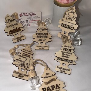 Può includere: Deodoranti per ambienti a forma di albero in legno con testo italiano, ciascuno con un piccolo flacone di vetro. Le frasi includono "Mio papà è il mio eroe" e "Ti amiamo papà". Un bouquet di rose rosa è sullo sfondo.